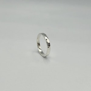 May include: A silver ring with a polished finish and a hammered texture on the outer surface. The band has a consistent width and a circular shape, reflecting light across its faceted metallic surface against a plain, neutral background.