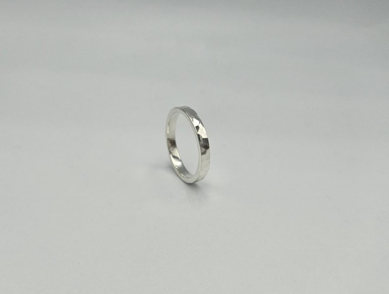 May include: A silver ring with a polished finish and a hammered texture on the outer surface. The band has a consistent width and a circular shape, reflecting light across its faceted metallic surface against a plain, neutral background.