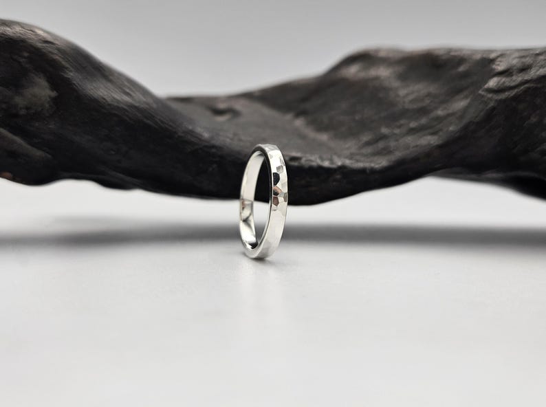 May include: A silver ring with a polished, hammered texture on the outer surface. The band has a rounded profile and a reflective metallic finish, showcasing small, irregular indentations across the entire exterior circumference of the metal.