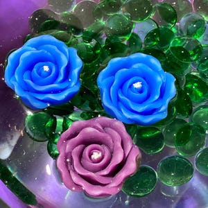 May include: Three rose-shaped candles in shades of blue and purple float among green glass beads in a purple bowl. The candles have white wicks and are surrounded by clear, round glass beads.