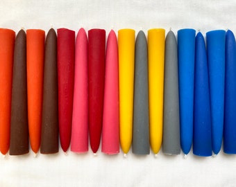 May include: A set of 18 taper candles in various colors, including orange, brown, red, pink, yellow, gray, and blue. The candles are arranged in a row on a white surface.