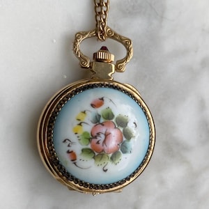 Op de afbeelding: Een goudkleurige, ronde hangerketting met een bloemmotief. De hanger heeft een lichtblauwe achtergrond met een handgeschilderde roze bloem en groene bladeren. De ketting heeft een delicate gouden ketting en een kleine rode edelsteen.