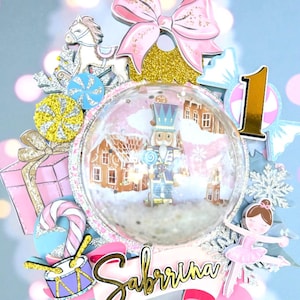 Könnte beinhalten: Festliche Tortenaufsatz mit einer Schneekugel, die einen Nussknacker und eine Dorflandschaft zeigt. Der Aufsatz enthält eine rosa Schleife, eine Ballerina, die Zahl 1 und den Namen "Sabrina". Die Farbpalette umfasst Rosa, Gold und Blau.