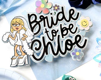May include: A cake topper featuring a cartoon illustration of a blonde person in a white dress, veil, and heels, with the text "Bride to be Chloe" in black script. The design includes iridescent flower accents and a clear plastic pick base.