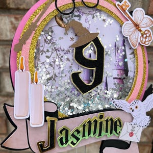Könnte beinhalten: Ein rosa-goldenes Harry-Potter-Themen-Tortenschild mit dem Namen "Jasmine". Der Aufsatz zeigt einen Zauberstab, eine Brille, einen Hut und einen Schlüssel. Die Zahl "9" ist prominent dargestellt, zusammen mit einer Schneeeule, die einen Brief hält. Zwei Kerzen sind ebenfalls enthalten.