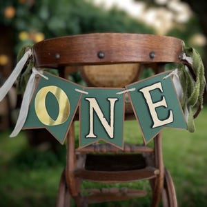 May include: A wooden high chair with a banner that says "ONE" in gold and white lettering. The banner is green with a pennant shape. The chair is decorated with white ribbons and green fabric.