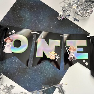 Op de afbeelding: Een zwarte banner met het woord "ONE" in grote, holografische letters, versierd met Star Wars-personages en sterren. De banner is ontworpen voor een verjaardagsfeest.