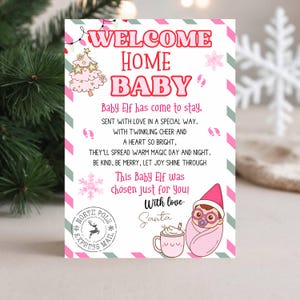 May include: A festive welcome home baby card with a pink and green border. The card features the text "WELCOME HOME BABY" and a poem about a baby elf. Illustrations include a Christmas tree, snowflake, baby elf, and a mug of hot chocolate.