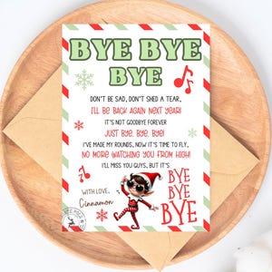May include: A festive card with the words "BYE BYE BYE" in green and red, featuring a cartoon elf illustration. The card includes a farewell message and is placed on a wooden plate with an envelope.