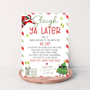 May include: A Christmas-themed greeting card with the words "Sleigh Ya Later" in green and red. The card features illustrations of Santa Claus, a Christmas tree, and a reindeer. The card is on a wooden tray.