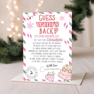 Puede incluir: Una tarjeta de felicitación navideña con el texto "¿Adivina quién está de vuelta?" en rosa y rojo. La tarjeta presenta un diseño festivo con copos de nieve, bastones de caramelo y una linda ilustración de un árbol rosa y una taza de chocolate caliente. La tarjeta está firmada "Cinnamon".