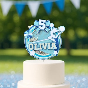 May include: A two-tiered white cake topped with a blue and white cake topper. The topper features the number 5, the name "OLIVIA", and a cheerleader silhouette. The background includes blue and white bunting and a blurred green backdrop.