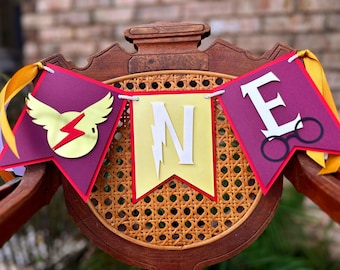 May include: A decorative banner spelling "ONE" across three pennants. The pennants are purple, yellow, and red, with gold and white accents. The banner features a lightning bolt, glasses, and a winged emblem, suitable for a birthday.