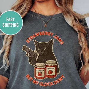 以下が含まれることがあります： 黒猫が銃を持っているダークグレーのTシャツ。赤字で「STRAWBERRY JAMS BUT MY GLOCK DON'T」と書かれています。イチゴジャムの瓶も2つ描かれています。このシャツはヴィンテージ風です。