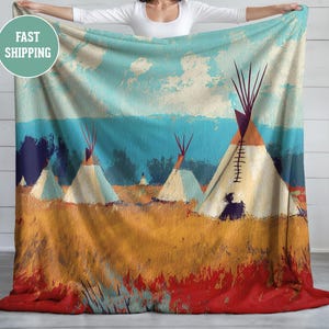 May include: A blanket featuring a landscape painting of teepees in a field. The artwork uses a palette of blues, oranges, and reds, with a sky and clouds in the background. The blanket is held up by a person.