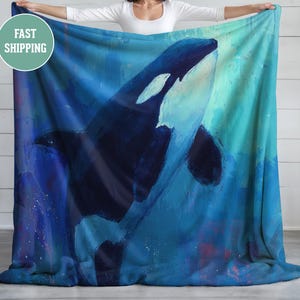 May include: A blue blanket featuring an artistic depiction of an orca whale. The whale is painted in shades of blue, white, and black, with a focus on the animal's form and the surrounding water. The blanket is held up by a person.