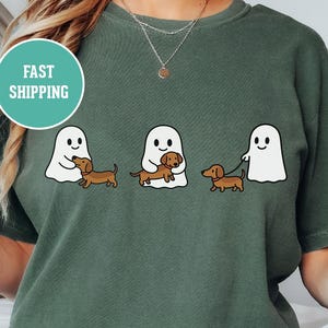 May include: Green t-shirt featuring a graphic of three white cartoon ghosts with brown dachshund dogs. One ghost holds a dog, one walks a dog on a leash, and one holds a dog. The text "FAST SHIPPING" is in a teal circle.