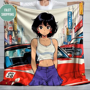 Op de afbeelding: Een zachte deken met een anime-stijl illustratie van een vrouw voor een rode auto in een stedelijke omgeving. De vrouw draagt een witte tanktop en een paarse short. De achtergrond bevat gebouwen en Japanse bewegwijzering.
