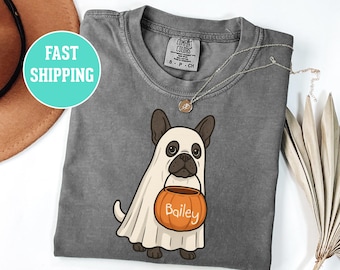 May include: A gray t-shirt featuring a cartoon French Bulldog dressed as a ghost, holding an orange pumpkin-shaped candy bucket with the name "Bailey" on it. The shirt has a round neckline and short sleeves. A silver necklace is draped on the shirt.