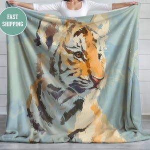 Op de afbeelding: Een zachte, lichtblauwe deken met een gedetailleerde schildering van een tijgergezicht. De tijger heeft oranje en bruine strepen, met een roze neus en expressieve ogen. De deken wordt omhoog gehouden, wat de levendige kleuren van het kunstwerk laat zien.