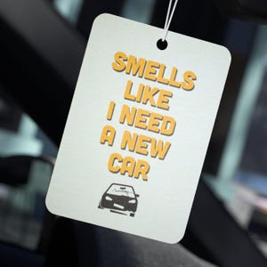 May include: White car air freshener with the text "Smells like I need a new car" in yellow letters. A black silhouette of a car is at the bottom of the air freshener.
