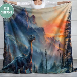 Può includere: Una coperta in pile blu e verde con una silhouette di dinosauro in primo piano e una catena montuosa con un tramonto sullo sfondo.
