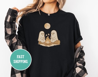 Puede incluir: Camiseta negra con un gráfico de dos fantasmas de dibujos animados asando malvaviscos sobre una fogata bajo una luna llena. El diseño incluye un tronco, llamas y una combinación de colores marrón claro.