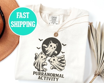 May include: Cream-coloured t-shirt with a graphic of three cartoon cats dressed as ghosts, with bats and a full moon. The text "PURRANORMAL ACTIVITY" is printed below the graphic. A gold necklace is draped on the shirt.