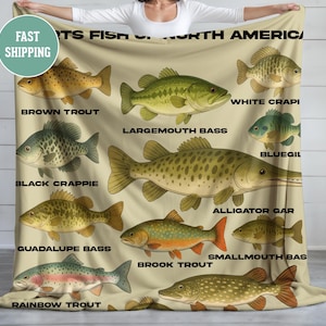 Puede incluir: Una manta de forro polar beige con ilustraciones de diferentes tipos de peces que se encuentran en América del Norte. Los peces están etiquetados con sus nombres, incluyendo trucha marrón, lubina de boca grande, crappie negro, crappie blanco, bluegill, gar de caimán, lubina de Guadalupe, lubina de boca pequeña, truite de arroyo y trucha arcoíris.