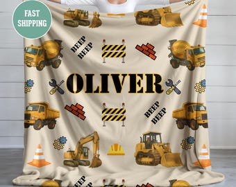 Op de afbeelding: Een beige deken met een bouwthema, met gele bouwmachines, pionnen en de naam "OLIVER". Het ontwerp bevat herhalende patronen van bouwgerelateerde afbeeldingen.