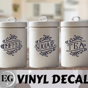 May include: Three white ceramic canisters with black vinyl decals. The canisters are labeled "Coffee", "Sugar", and "Tea".