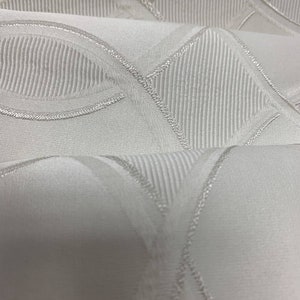 May include: Close-up of a white fabric with a textured design. The fabric features a repeating pattern of curved lines and diagonal stripes, all outlined with a subtle, silvery thread. The folds in the fabric create shadows, adding depth to the design.