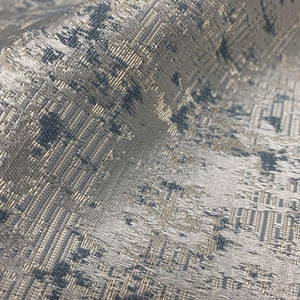 May include: Close-up of a textured fabric with a woven pattern. The fabric has a mix of silver, gold, and blue-gray tones, creating a shimmering effect. The weave is detailed, with a slight sheen, suggesting a luxurious textile.