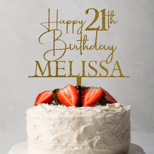 May include: A white frosted cake with sliced strawberries on top, presented on a white cake stand. A gold glitter cake topper reads "Happy 21st Birthday MELISSA".