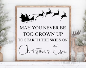 May include: A white wooden sign with black text that reads "MAY YOU NEVER BE TOO GROWN UP TO SEARCH THE SKIES ON Christmas Eve". The sign features a black silhouette of Santa Claus in his sleigh with reindeer flying through the air.