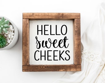 May include: A wooden framed sign with a white background and black text that reads "HELLO sweet CHEEKS".
