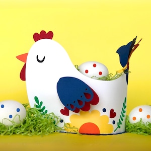 Puede incluir: Una cesta de papel blanca con forma de gallina con cresta roja, pico naranja y detalles de alas azules y rojas. La cesta contiene huevos blancos con lunares rojos, metidos en hierba de papel verde, sobre un fondo amarillo.