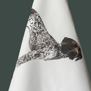 May include: A white kitchen towel featuring a detailed illustration of a German Shorthaired Pointer dog. The dog has a brown head and a white coat with brown ticking and spots. The towel is hanging from a hook, displaying the full canine graphic.