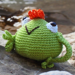 Può includere: Peluche di rana verde all'uncinetto con capelli in lana arancione e occhi in lana bianca. La rana è seduta su uno scoglio.
