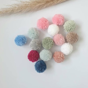 Könnte beinhalten: Sortiment an bunten Pompons in verschiedenen Farbtönen von Rosa, Blau, Grün, Grau und Rot. Die Pompons sind aus Garn gefertigt und weich und flauschig.
