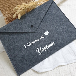 Könnte beinhalten: Graue Filztasche mit einem weißen Herz und dem Text "Hebamme mit Yasemin" in weiß.