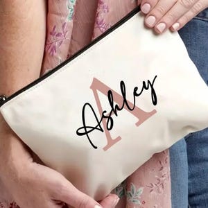 Könnte beinhalten: Cremefarbene Canvas-Tasche mit schwarzem Reißverschluss. Auf der Tasche steht der Name "Ashley" in schwarzer Schrift, mit einem großen rosa Buchstaben "A" im Hintergrund. Die Tasche wird von einer Person gehalten.