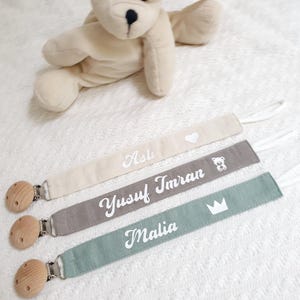 Könnte beinhalten: Drei personalisierte Schnullerketten mit Holzclips. Die Ketten sind beige, grau und grün. Die Namen "Asli", "Yusuf Imran" und "Malia" sind auf die Ketten gestickt. Die beige Kette hat ein weißes Herz, die graue Kette einen weißen Bären und die grüne Kette eine weiße Krone.