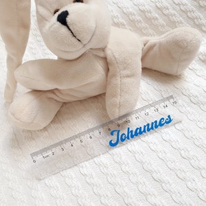 Könnte beinhalten: Ein durchsichtiges Plastiklineal mit einer blauen Aufschrift "Johannes" und Markierungen in Zentimetern von 0 bis 15. Das Lineal liegt auf einer weißen gestrickten Oberfläche mit einem Plüsch-Hasenspielzeug.
