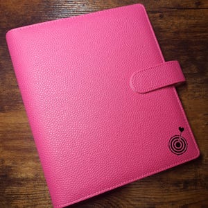 May include: A bright pink, textured leather-like planner with a magnetic closure. The planner has a black graphic of a heart and a target on the lower right corner. The planner is closed and sits on a wooden surface.