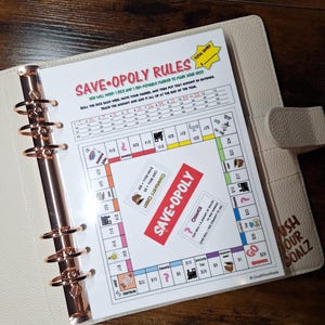 May include: A planner with a "Save-Opoly Rules" game board insert. The game board has spaces for tracking savings, with text that reads "Crush Your Goalz" on the planner cover. The planner is open to the game board.
