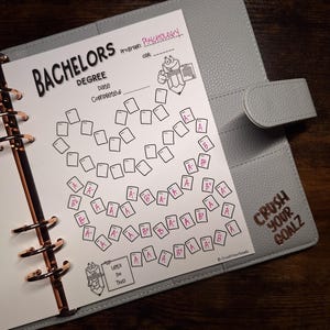 Op de afbeelding: Een lichtgrijze planner met roségouden ringen met een witte papieren tracker voor een bachelordiploma in de psychologie. De pagina bevat een hartvormig raster met handgeschreven cijfers, een potloodfiguurtje en de tekst Crush Your Goalz.