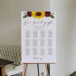 Pode incluir: Um cartaz branco de plano de mesas de casamento num cavalete de madeira com um arranjo floral em aguarela de um girassol e rosas vermelho-escuras no topo. O texto diz Our favorite people, Ashley + Adam, 21 de setembro 2022, mesas e Thank you.