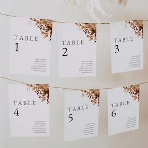 Puede incluir: Tarjetas blancas con números de mesa, numeradas del 1 al 6, suspendidas de una cuerda. Cada tarjeta presenta un diseño floral en la esquina superior derecha y los nombres de los invitados debajo. Las tarjetas se sujetan con clips dorados. El texto "TABLE" está en la parte superior de cada tarjeta.