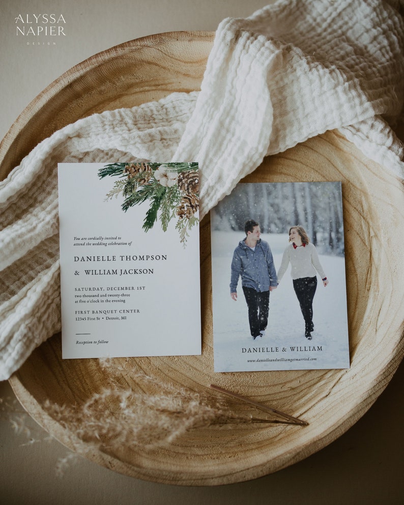 Puede incluir: Juego de papeler&iacute;a de boda con una invitaci&oacute;n y una tarjeta fotogr&aacute;fica. La invitaci&oacute;n presenta texto y un dise&ntilde;o bot&aacute;nico con pi&ntilde;as y vegetaci&oacute;n. La tarjeta fotogr&aacute;fica muestra a una pareja en la nieve. Ambos est&aacute;n en una bandeja de madera.